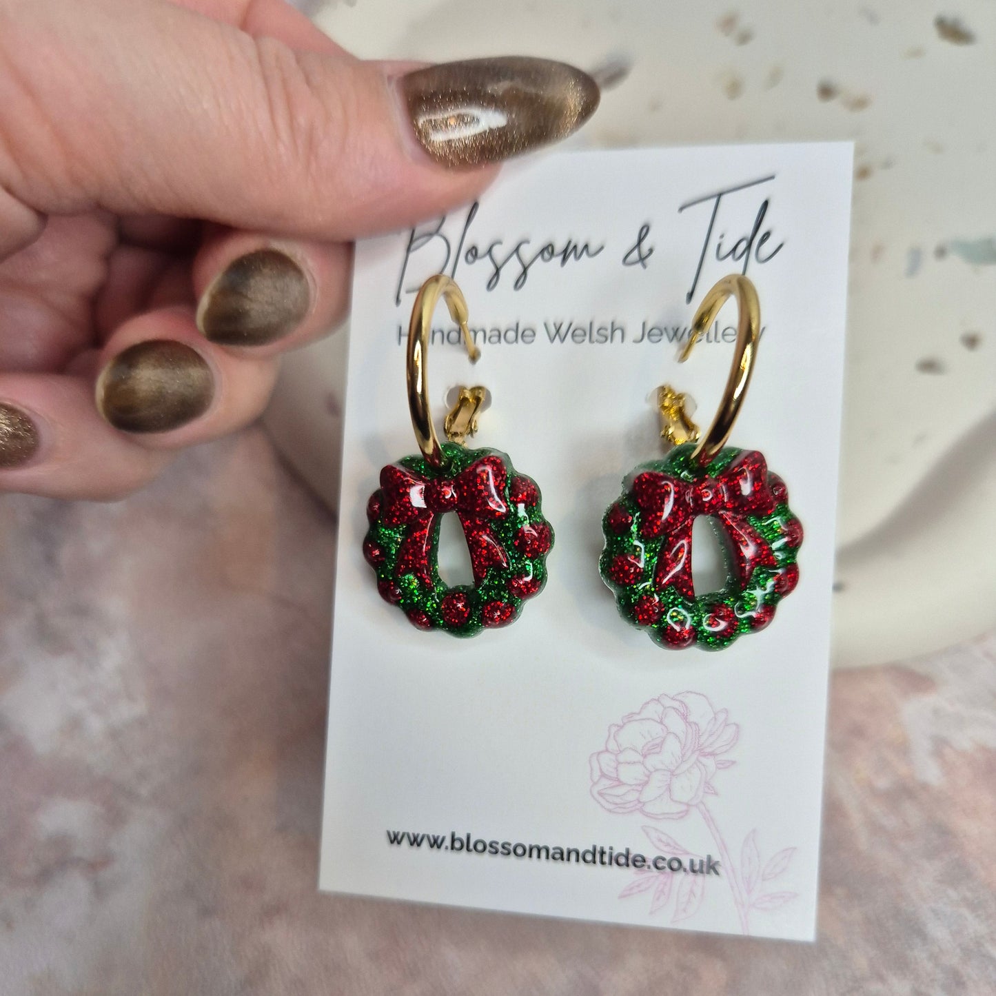 Red and green wreath earrings on a display card held by a hand with gold nail polish.