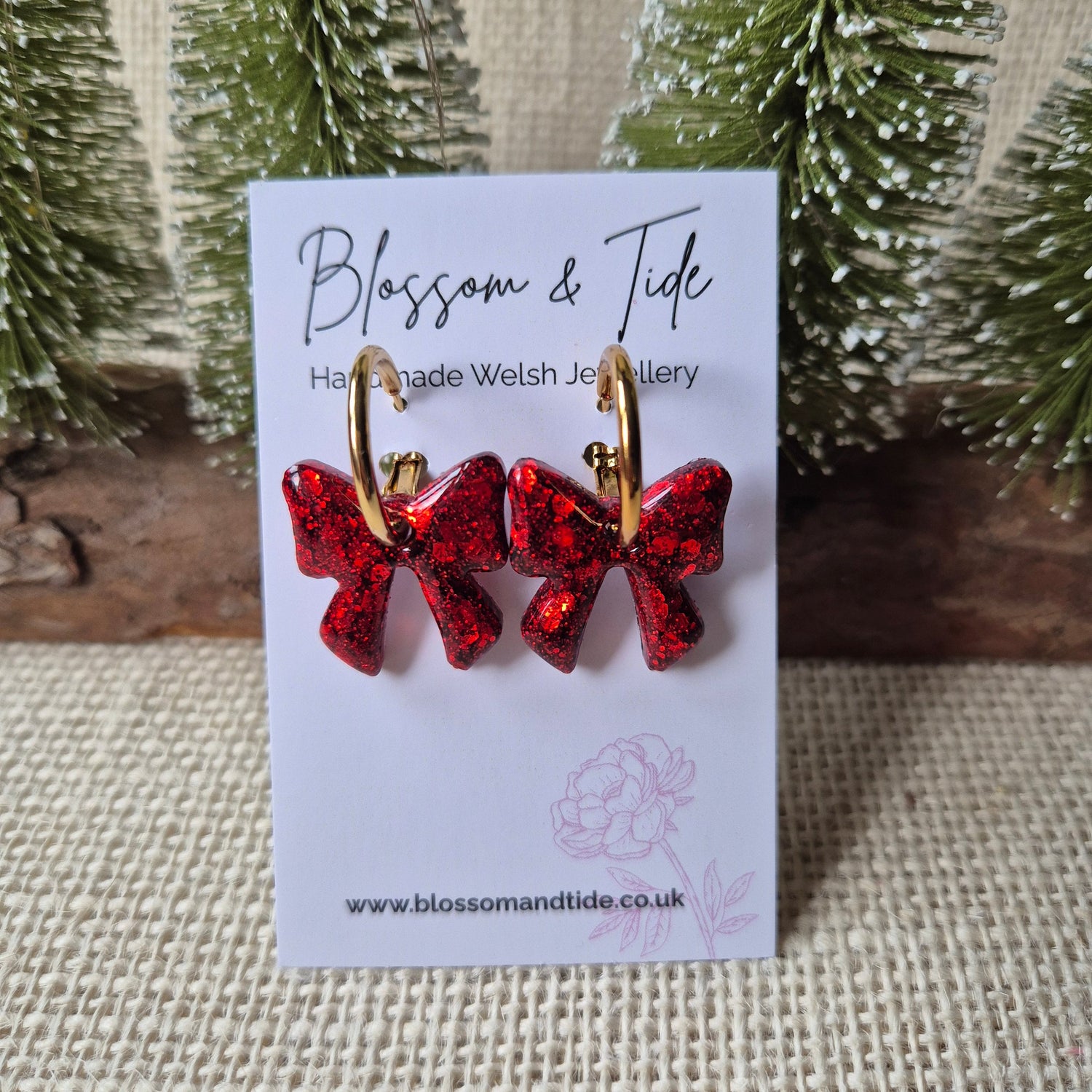 red glitter bow earrings on a gold hoop on a blossom and tide card, against a neutral background and christmas trees