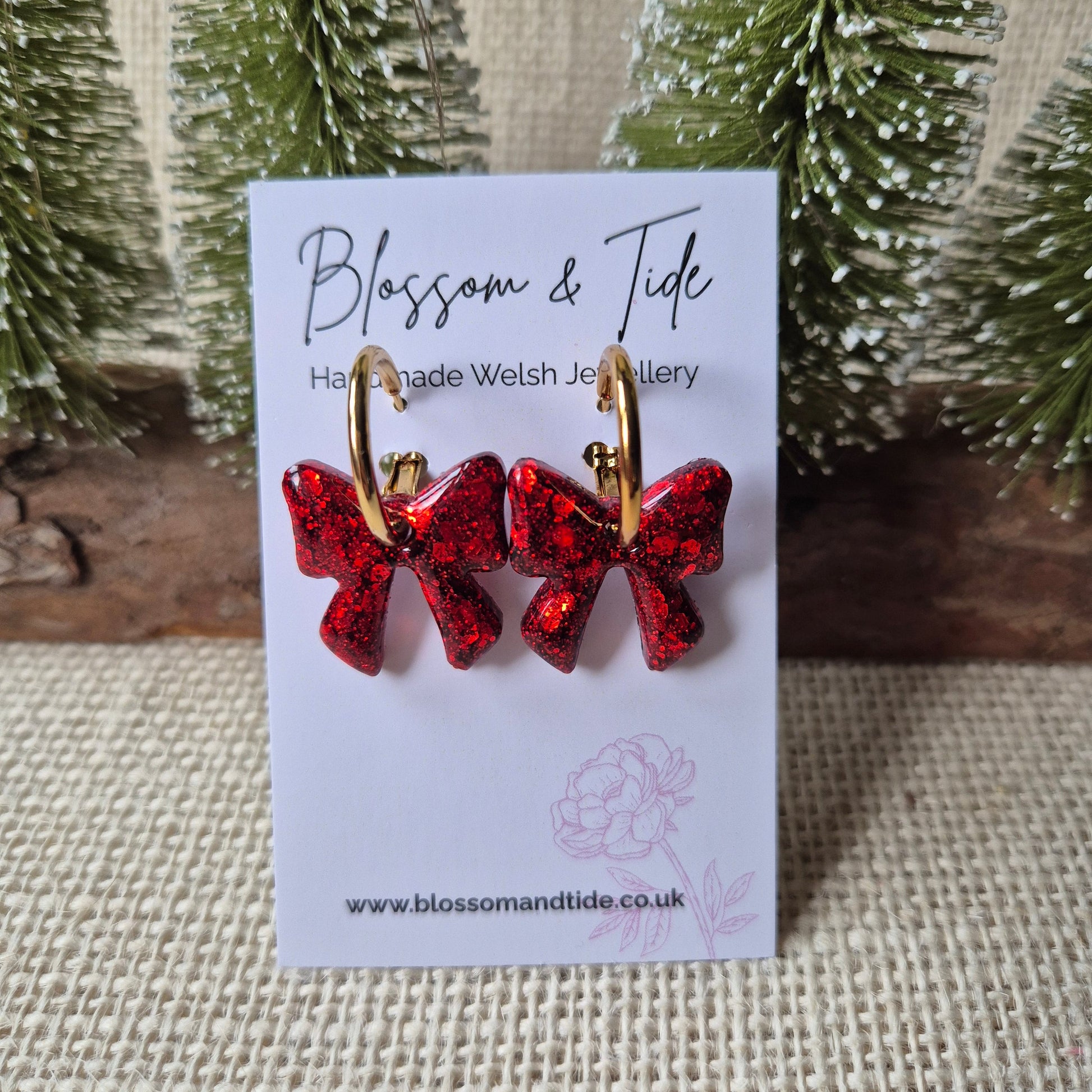 red glitter bow earrings on a gold hoop on a blossom and tide card, against a neutral background and christmas trees