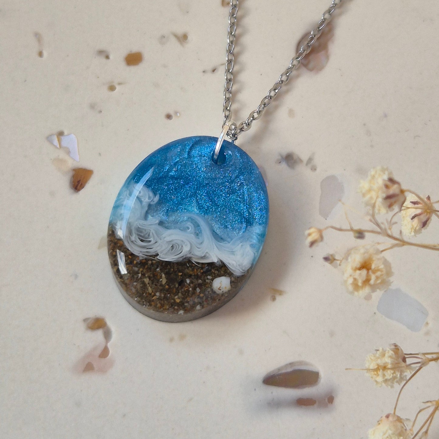 Necklace with a oval pendant featuring sand and sea design on a light background with small flowers and stones.