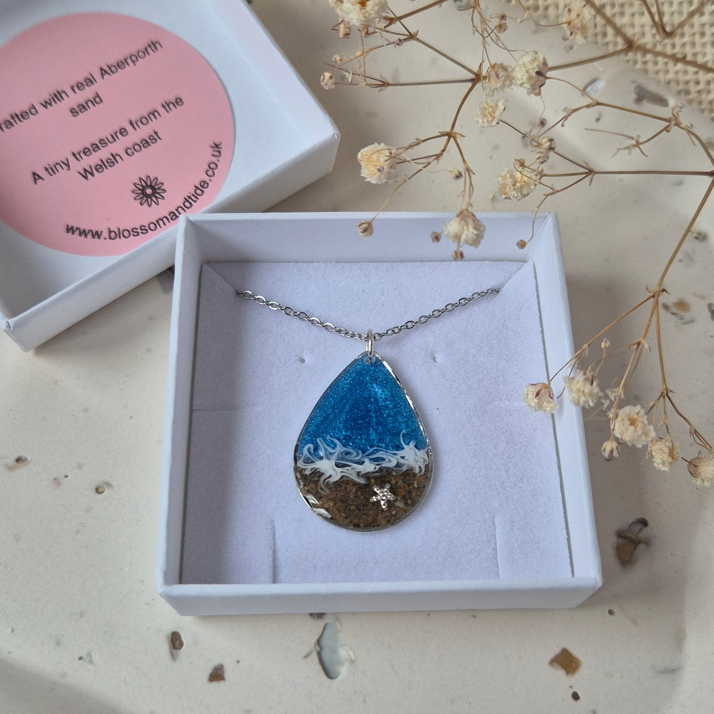 Necklace with a blue sea and sand pendant in a white box on a textured surface with dried flowers.