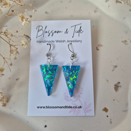 Blue triangle-shaped earrings on a 'Blossom & Tide' card with dried flowers in the background.